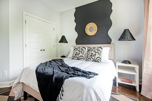 Quarto com cama, duas luminárias de cabeceira, mesa de cabeceira com livros e um objeto decorativo na parede.