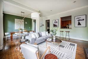 Sala de estar moderna com um sofá cinza, uma mesa de centro e um tapete estampado. Há uma área de jantar com uma mesa e cadeiras brancas, e uma cozinha visível por uma porta aberta.