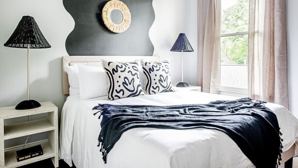 A bedroom with a bed, two lamps, a shelf with books, a wall-mounted mirror, and a window with curtains.
