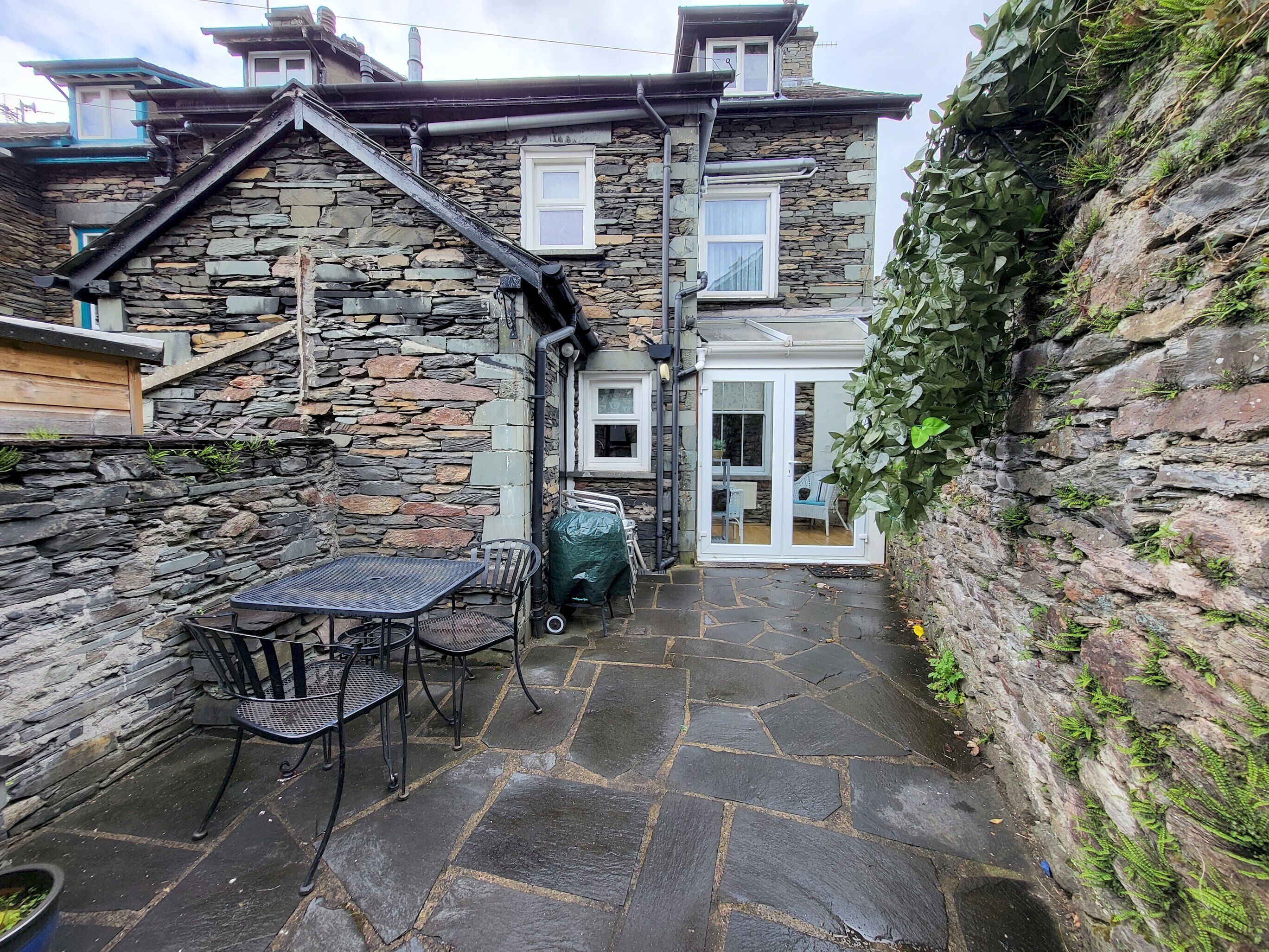 Ein Steinhaus mit einer Terrasse, die einen Tisch, Stühle, einen überdachten Grill und einen kleinen Garten beherbergt.