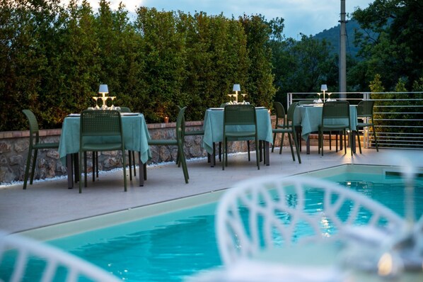 A poolside area with tables and chairs, a stone wall, and a view of trees and hills.