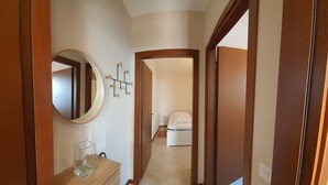 Un couloir mène à une chambre qui contient un lit, une commode, un miroir et un luminaire fixé au mur.