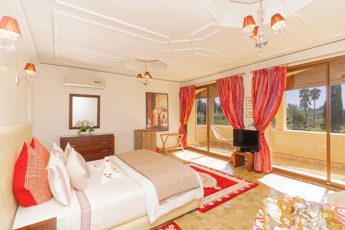 A spacious bedroom with a large bed, a television, and a balcony with red curtains.