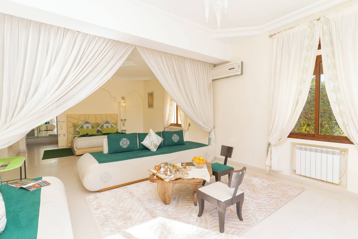 A spacious living room with a large sofa, a coffee table, and a dining area. There is a window with curtains and a view of greenery outside.