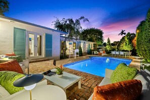 Un área junto a la piscina con un patio de ladrillo, mobiliario exterior y un pequeño edificio con persianas azules.