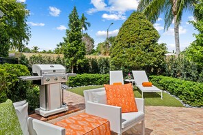 A patio with a grill, white chairs, and orange cushions.