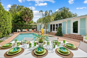 Un área de comedor junto a la piscina, con una mesa dispuesta para seis personas, rodeada de exuberante vegetación y una piscina.