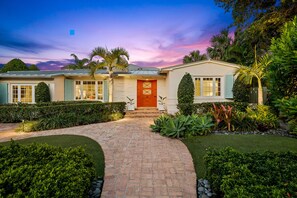 Una casa de dos pisos con un sendero de ladrillo que conduce a la entrada, rodeada de setos cuidados y plantas tropicales.