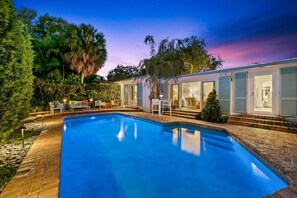 Une piscine avec un espace patio, entourée d’une maison dotée de portes coulissantes et d’un chemin en briques.