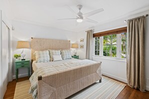 Une chambre à coucher comprenant un lit, des tables de chevet, un ventilateur de plafond, une fenêtre avec des rideaux et une vue sur la verdure à l’extérieur.