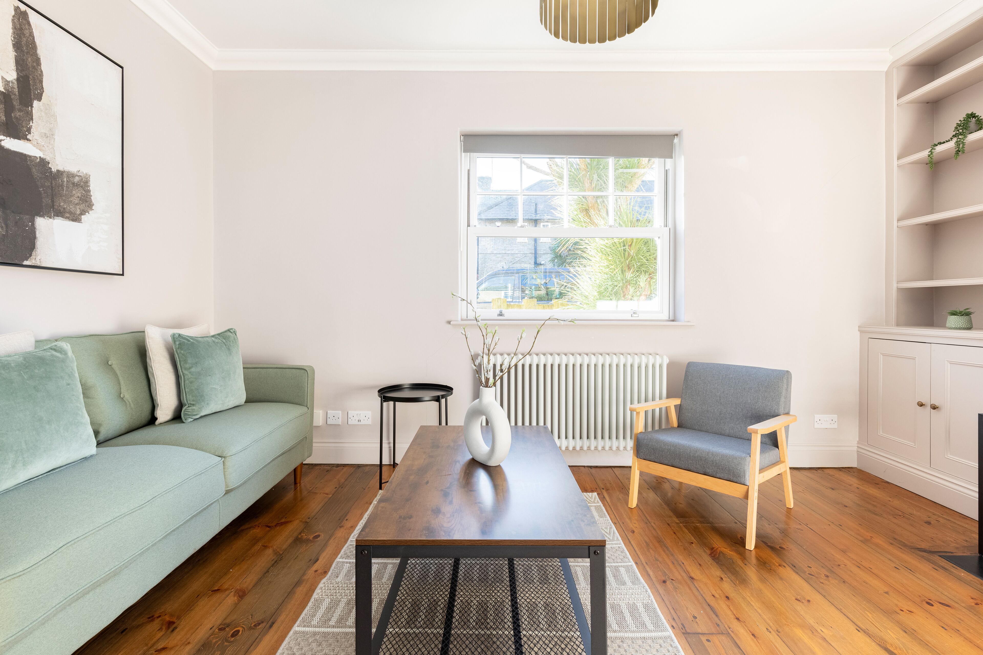 A living room with a light green sofa, a grey armchair, a wooden coffee table, a white vase, and a window with a view of houses.