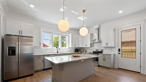 Une cuisine moderne avec des meubles blancs, un réfrigérateur en acier inoxydable, un grand îlot central et des luminaires suspendus.