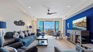 A modern living room with a sofa, armchairs, a coffee table, and a TV. There is a ceiling fan, a large window with a view of the ocean, and a bookshelf. - Oceanfront/Ground Floor Unit/A/C/Parking/Guest Favorite (La Jolla)