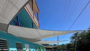 A covered outdoor area with a blue building, a shaded seating area, and a metal fence.