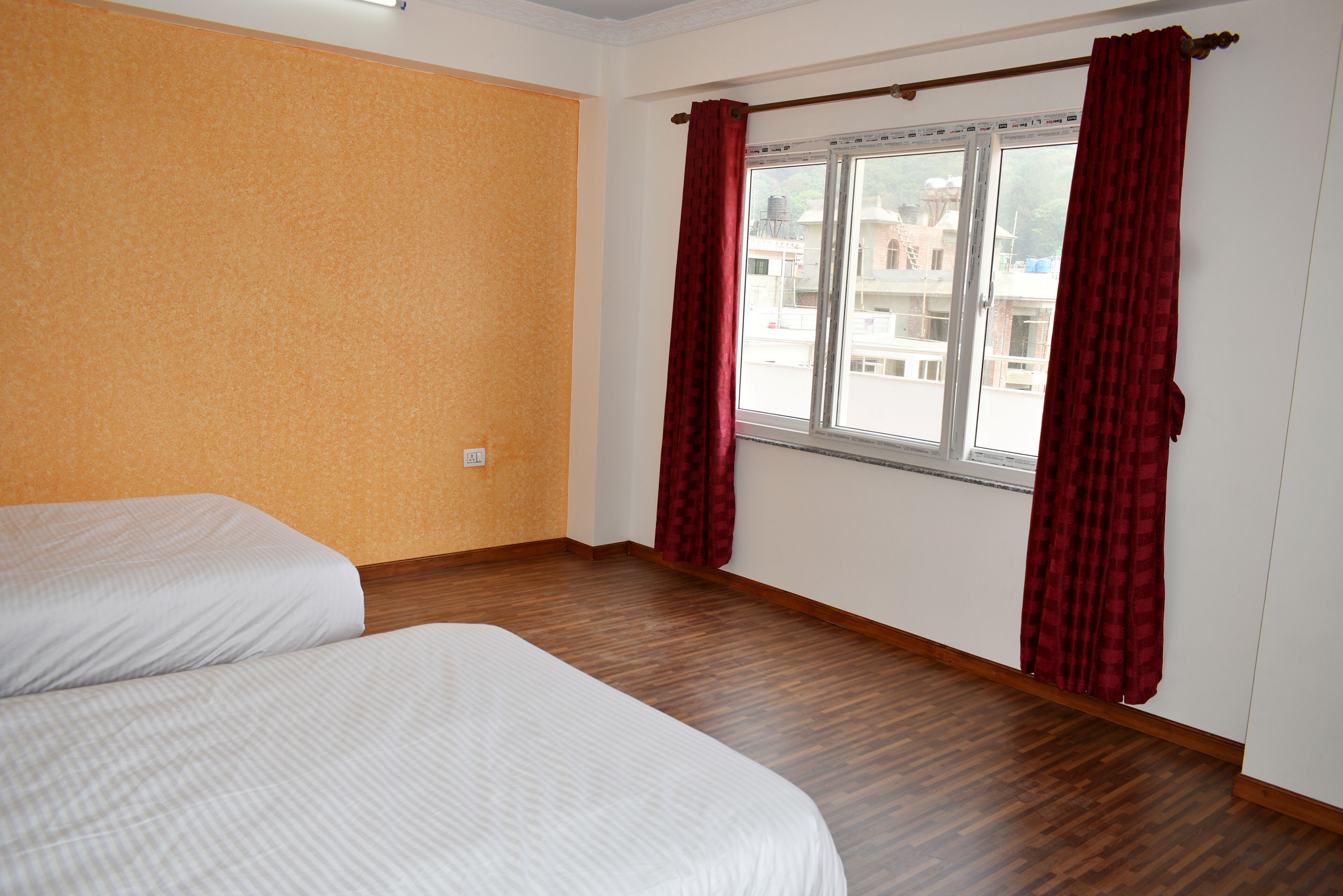 A room with two beds, a window with red curtains, and a view of buildings outside.