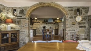 A rustic dining area with a wooden table and chairs, a stone wall, and a kitchen in the background.
