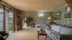A rustic living room with stone walls, a grey sofa, a wooden coffee table, and a view through an open door.