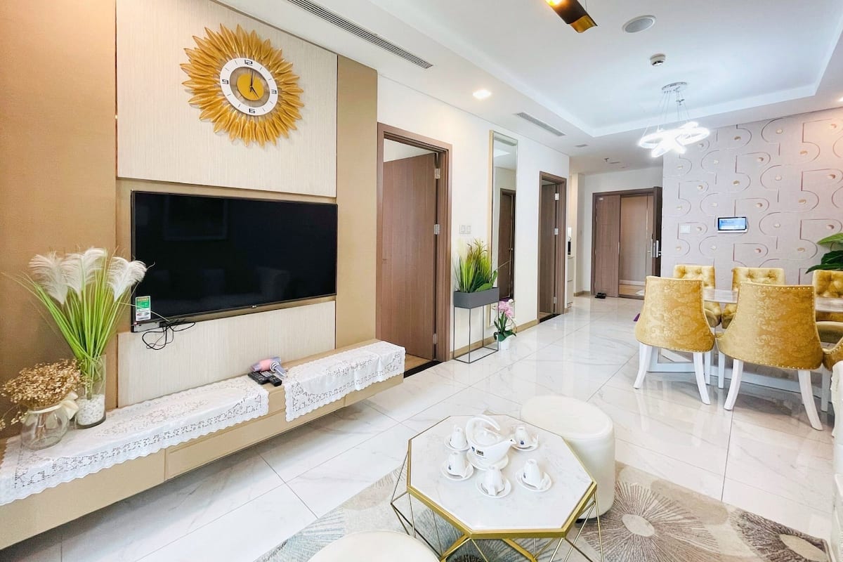 A modern living room with a large TV, a decorative sunburst wall piece, a seating area with a white table and chairs, and a dining area with a white table and chairs.