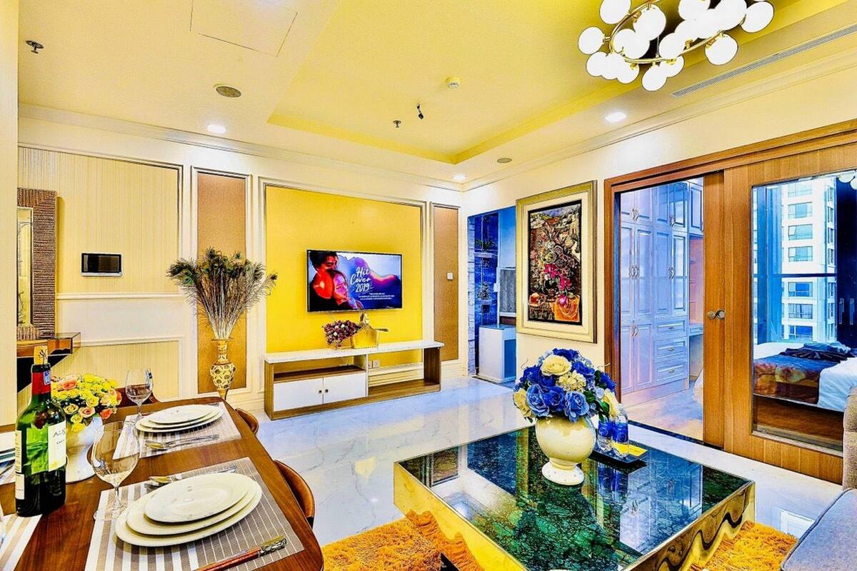 A modern living room with a yellow accent wall, a flat-screen TV, a dining area with a table set for two, and a large window offering a city view.