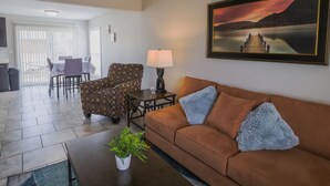 Ein Wohnzimmer mit braunem Sofa, einem gemusterten Sessel, einem Esstisch mit Stühlen, einer Lampe und einem gerahmten Bild an der Wand.