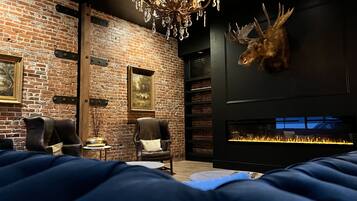 Une pièce avec un mur en briques, un lustre, une tête de cerf fixée au mur, une cheminée et deux fauteuils en cuir.