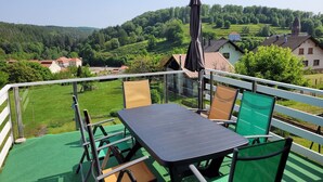 Uma varanda com mesa e cadeiras, com vista para uma paisagem verde com casas e uma colina.