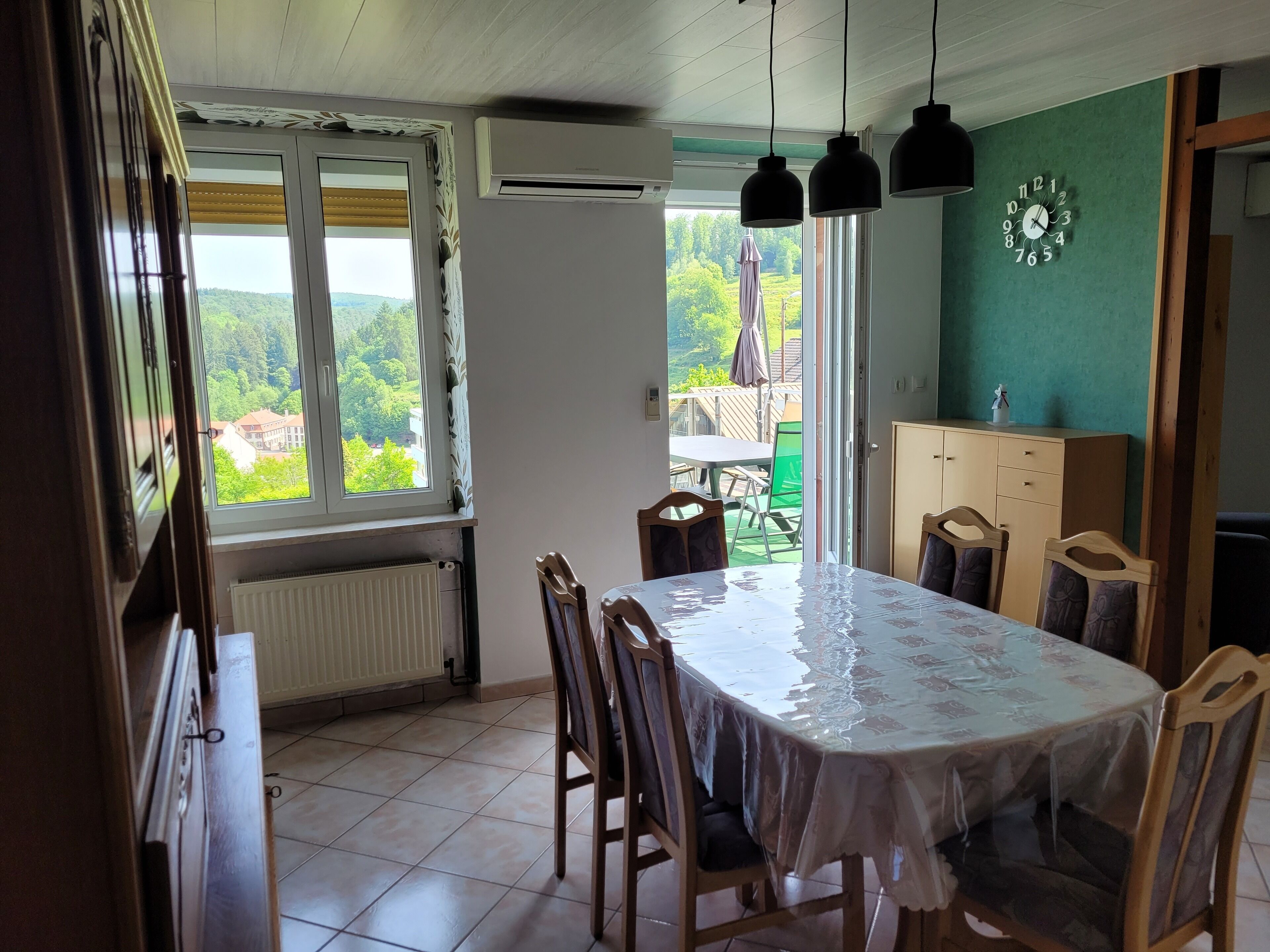 Een eetruimte met een tafel en stoelen, een uitzicht door een raam, en een klok aan de muur.