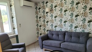 Uma sala de estar com um sofá cinza, uma poltrona cinza e uma pequena mesa preta com um centro de mesa trançado. O quarto tem um papel de parede estampado e uma janela com vista externa.