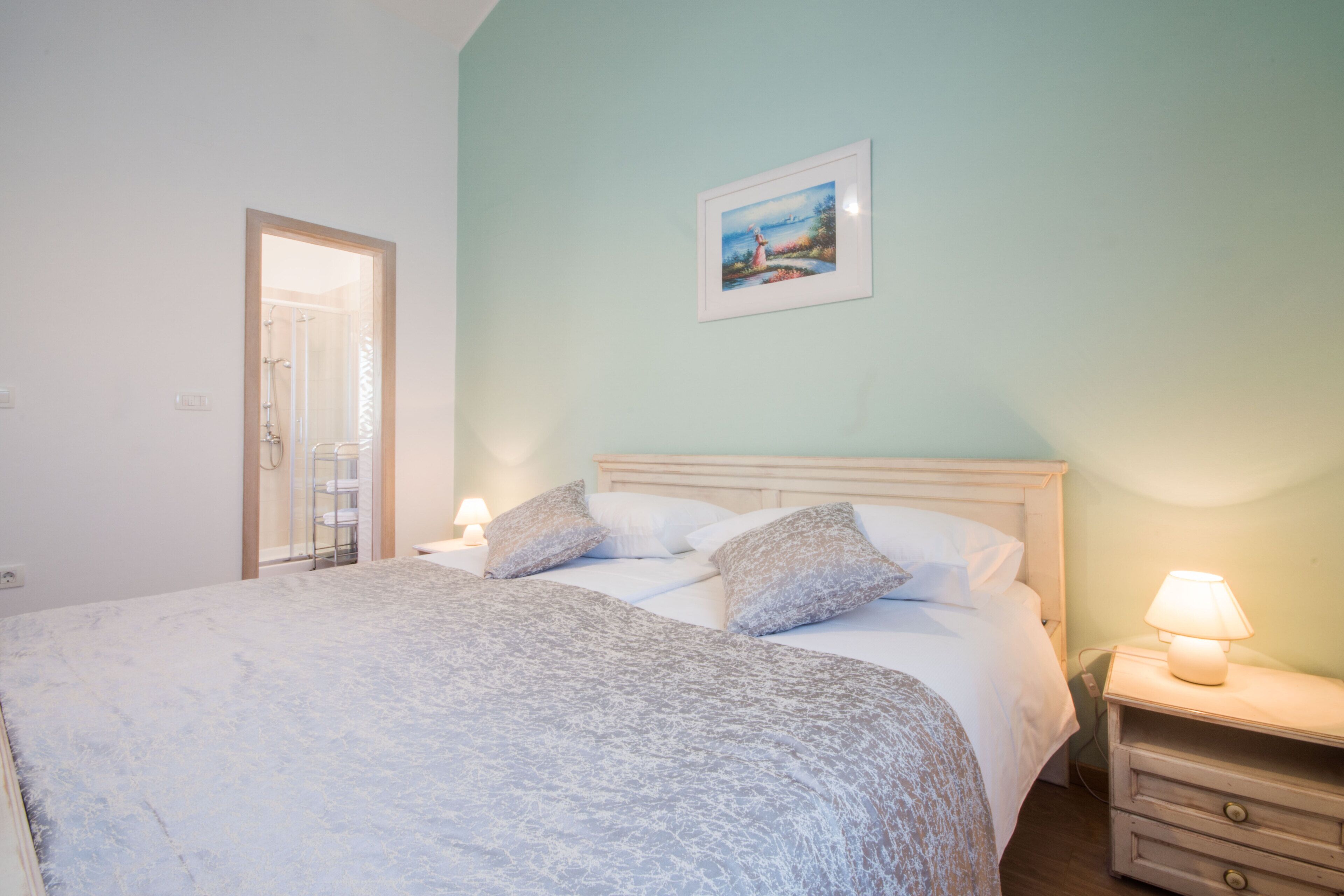 Quarto com cama, mesas de cabeceira, um criado-mudo, um quadro na parede e uma porta para o banheiro.