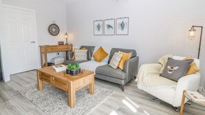 A modern living room with a grey sofa, a wooden coffee table, and a white armchair. There is a small wooden side table with a lamp, and a wall clock.