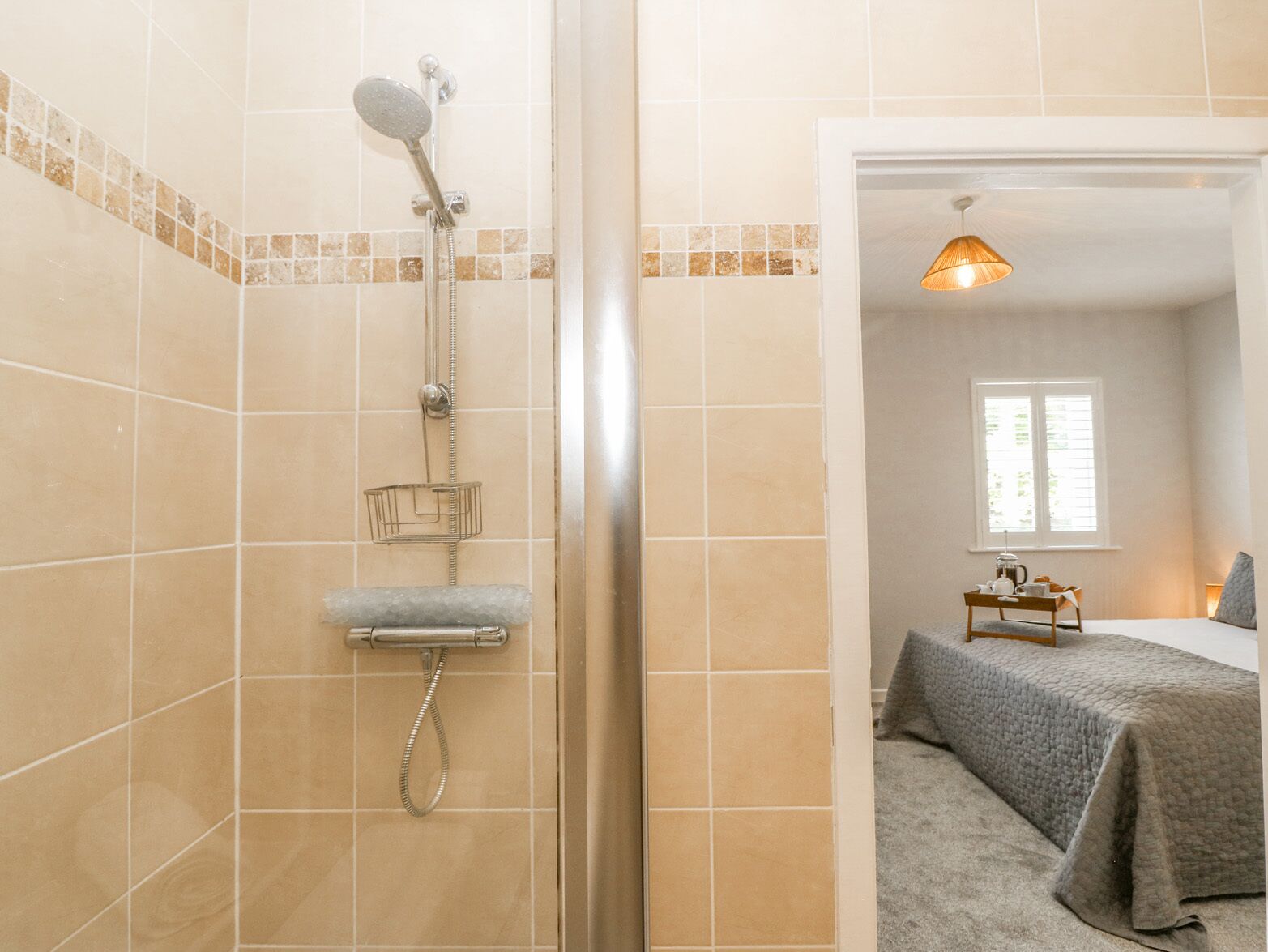 A bathroom with a shower, a towel rack, and a glass door leading to a bedroom with a bed and a window.
