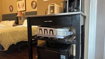 A hotel room with a bed, a nightstand, a lamp, a microwave, a coffee maker, and a small shelf with laundry baskets.