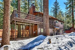 Un chalet rustique doté d’une terrasse en bois et d’une cheminée, entouré de grands arbres et de neige.