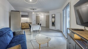 A modern living room with a blue sofa, a white coffee table, and a TV on a wooden stand. There is a dining area with a table and chairs, and a kitchen in the background. - View Point Semnoz 4* - OG IMMO (Annecy)