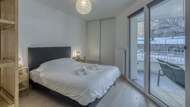 A modern bedroom with a large bed, a bedside table with a lamp, a sliding glass door to a balcony, and a view of a snowy landscape. - View Point Semnoz 4* - OG IMMO (Annecy)