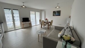 Una sala de estar moderna con sofá, una mesa de comedor con sillas y televisión. Se observan grandes ventanales y un balcón.