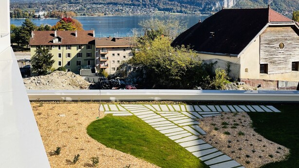 Vista de un lago con montañas al fondo, una zona de césped con un camino y edificios en primer plano.