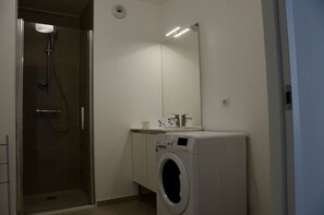 Un baño con ducha envuelta en vidrio, lavadora y espejo sobre el lavabo.
