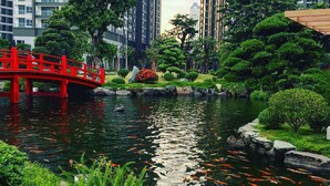 A serene water garden with a red bridge, koi fish, and lush greenery, surrounded by tall buildings. - DongDong LUX APT - Vinhomes Central Park (Ho Chi Minh City)
