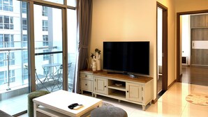 A modern living room with a large flat-screen TV, a white coffee table, and a grey ottoman. - DongDong LUX APT - Vinhomes Central Park (Ho Chi Minh City)