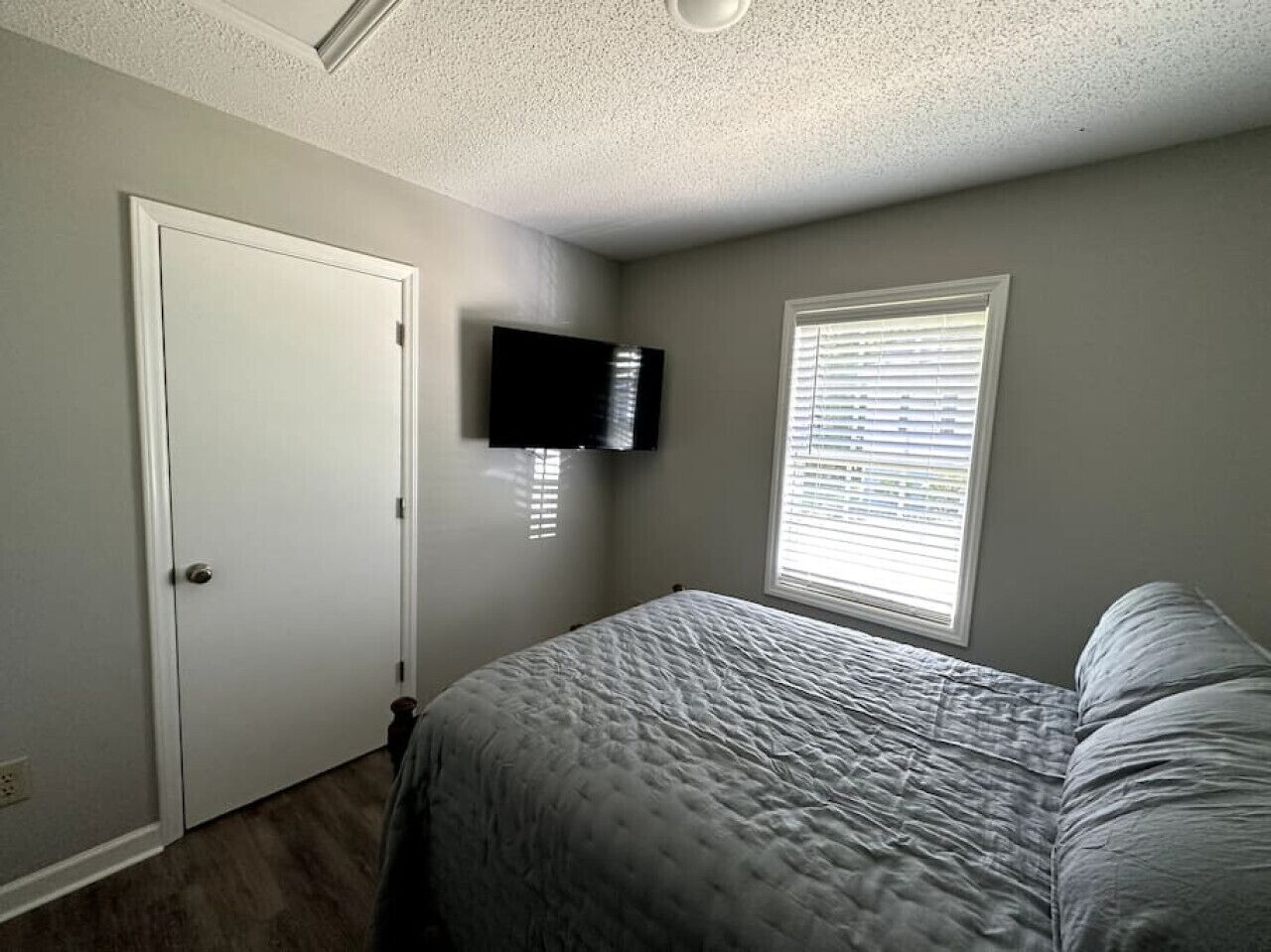 A bedroom with a bed, a television mounted on the wall, a door, and a window with blinds.