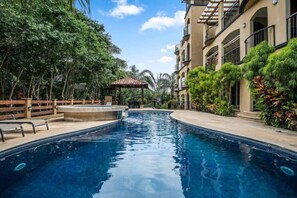 Una piscina con cubierta de madera y sillas de descanso, rodeada de abundante vegetación y un edificio de varios pisos con balcones.