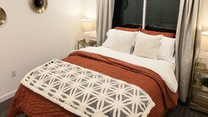 A neatly made bed with a patterned throw, two bedside lamps, and a window with curtains. - Downtown Stay at the Blue Tiny Twin (Roseburg)