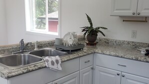 A kitchen with white cabinets, granite countertops, a sink, a window, and a potted plant. - Downtown Stay at the Blue Tiny Twin (Roseburg)