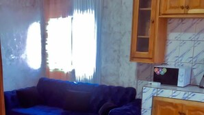 A living room with a blue sofa, a wooden coffee table, a kitchen area with cabinets and a microwave, and a tiled floor. - LA DIVA (Douala)