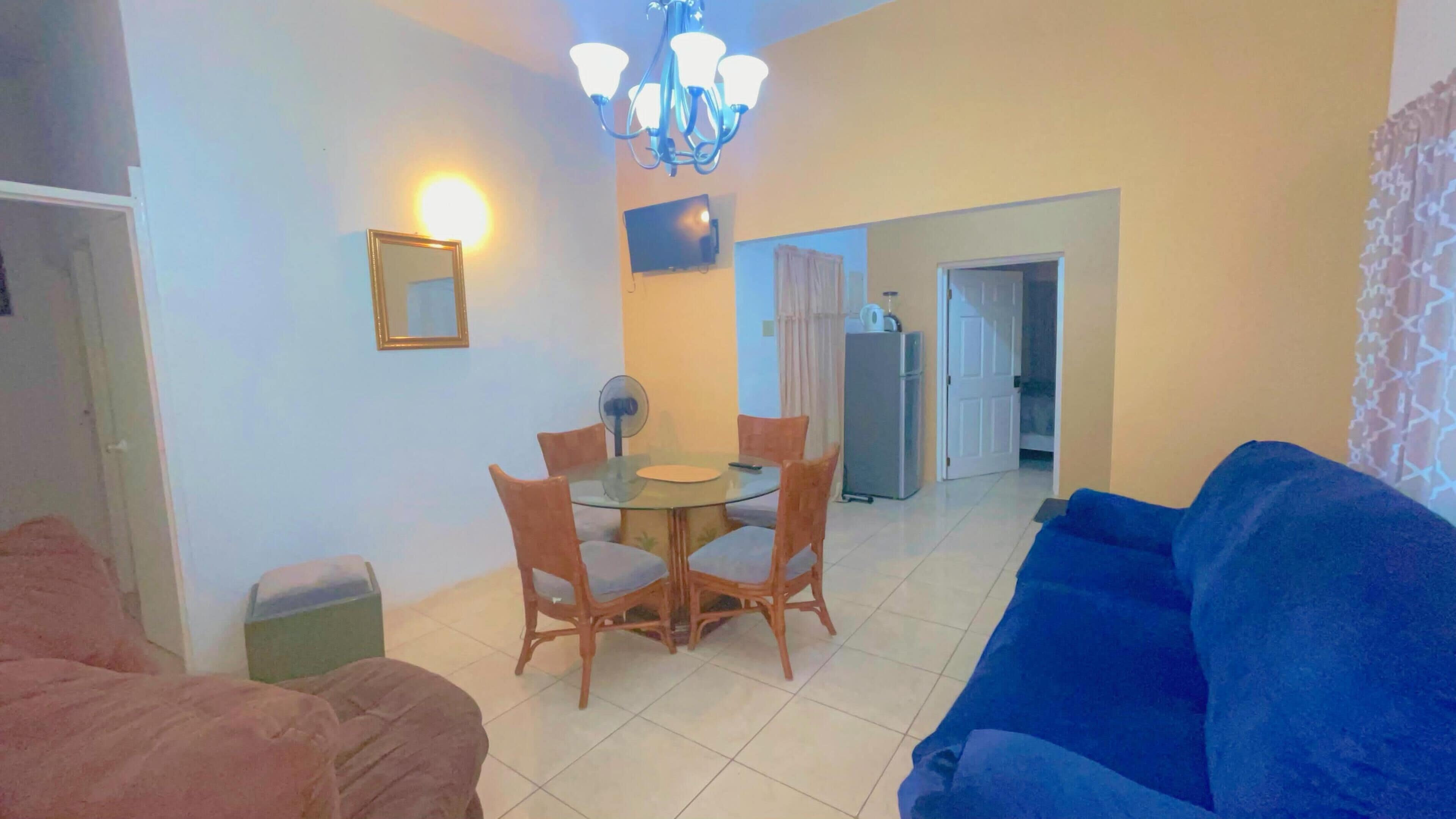 A living room with a blue sofa, a dining table with chairs, a chandelier, a TV, and a refrigerator in the background.