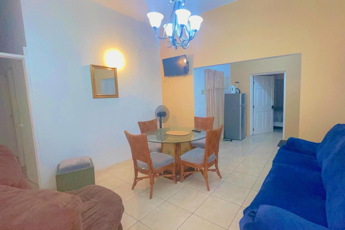 A living room with a blue sofa, a dining table with chairs, a chandelier, a TV, and a refrigerator in the background.