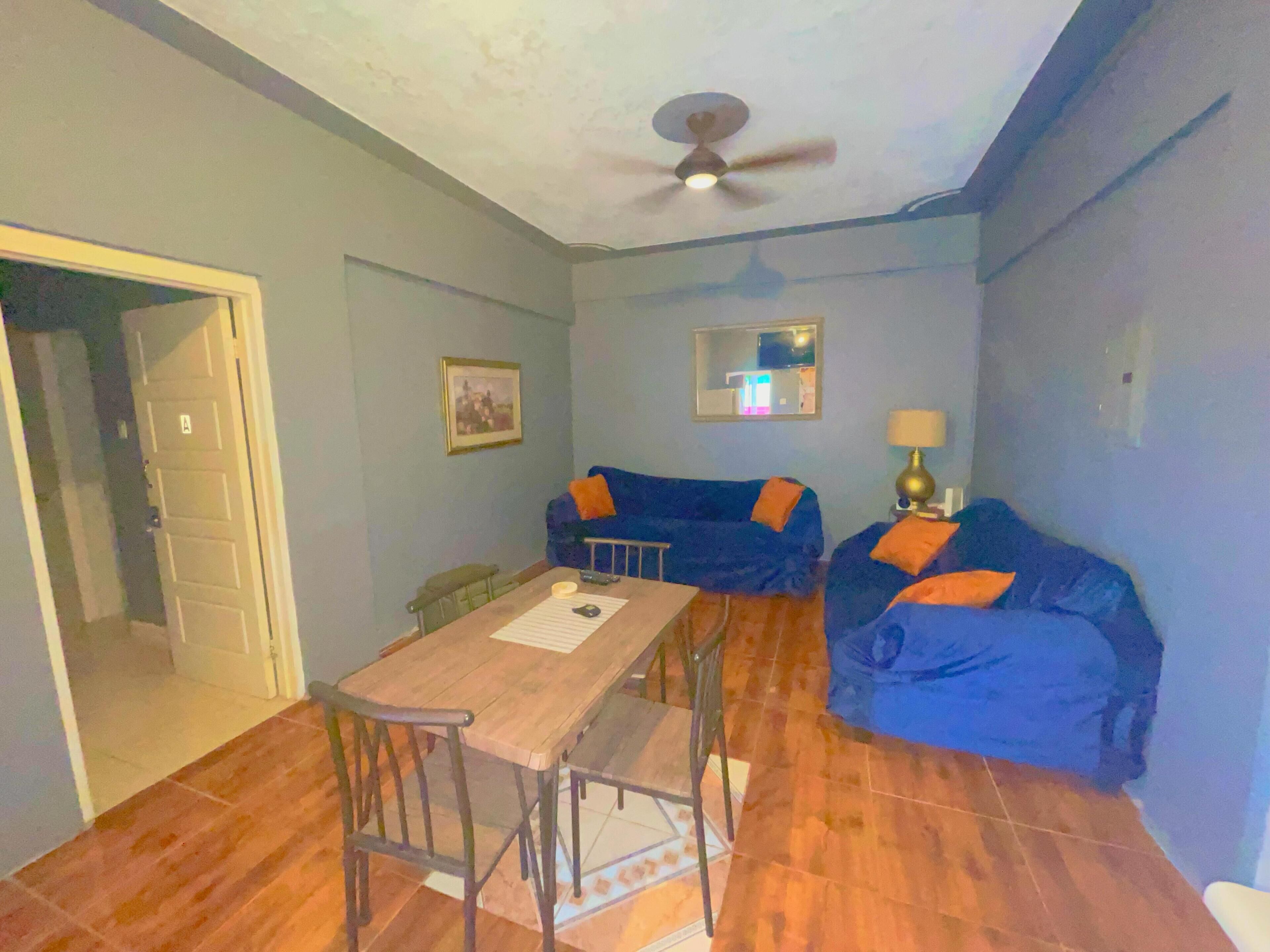 A living room with a wooden table and two blue sofas with orange cushions, a ceiling fan, and a framed picture on the wall.
