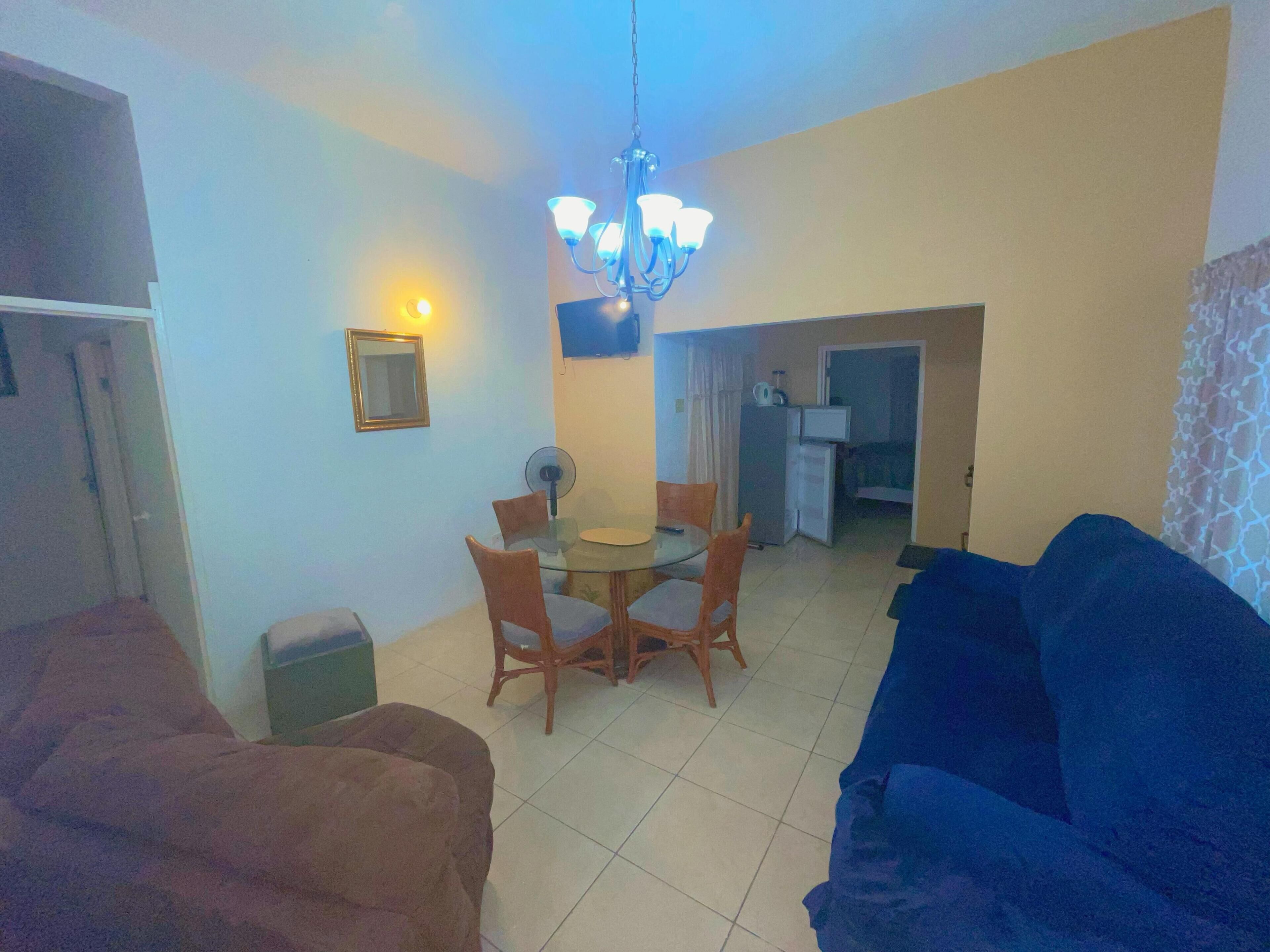 A living room with a blue sofa, a brown sofa, a dining table with chairs, a chandelier, and a kitchen area in the background.