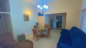 Una sala de estar con un sofá azul, un sofá marrón, una mesa de comedor con sillas, una araña y una cocina al fondo.